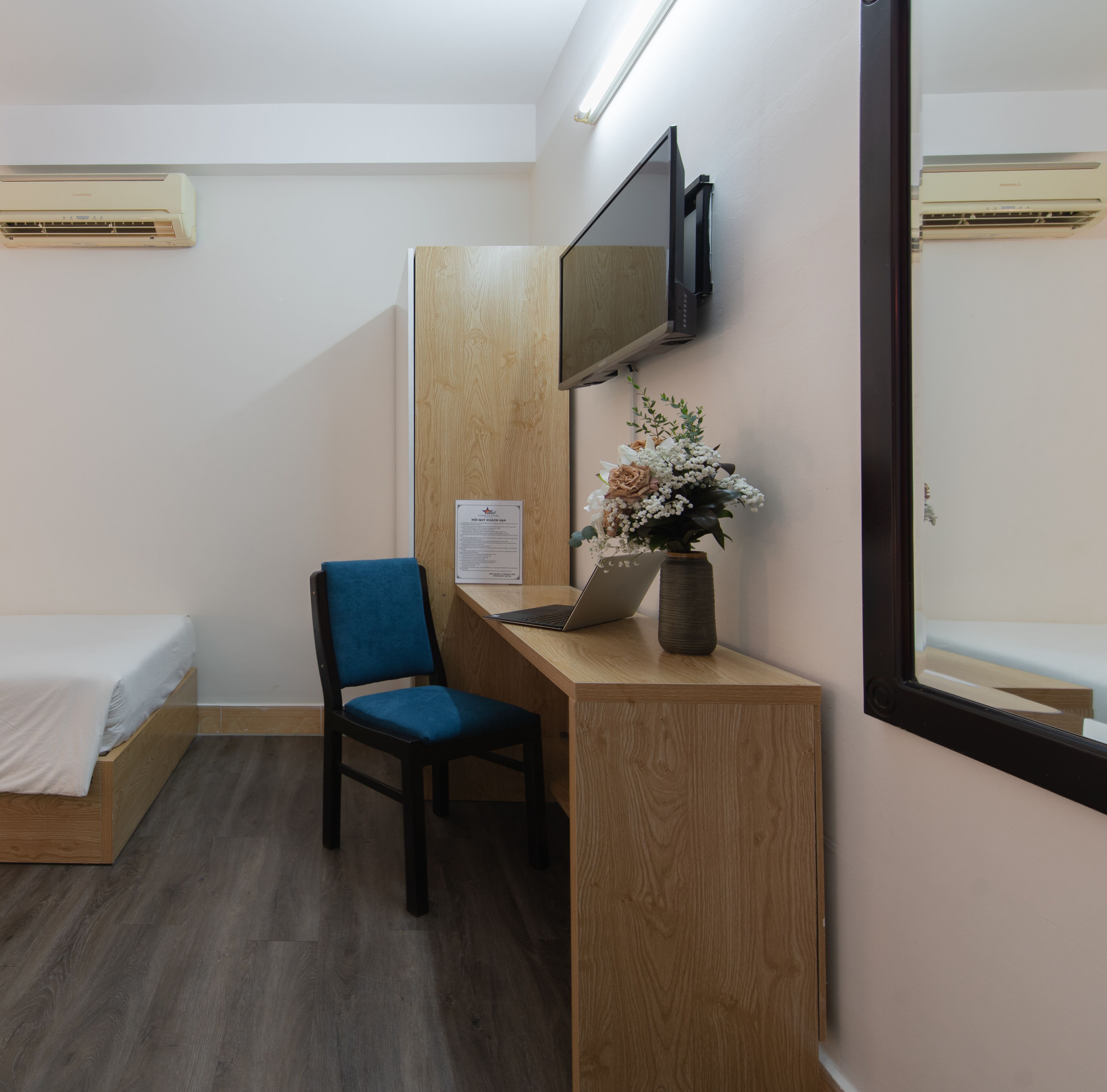 A hotel room with a bed, a desk with a chair, a laptop, a vase with flowers, a mirror, and an air conditioner.