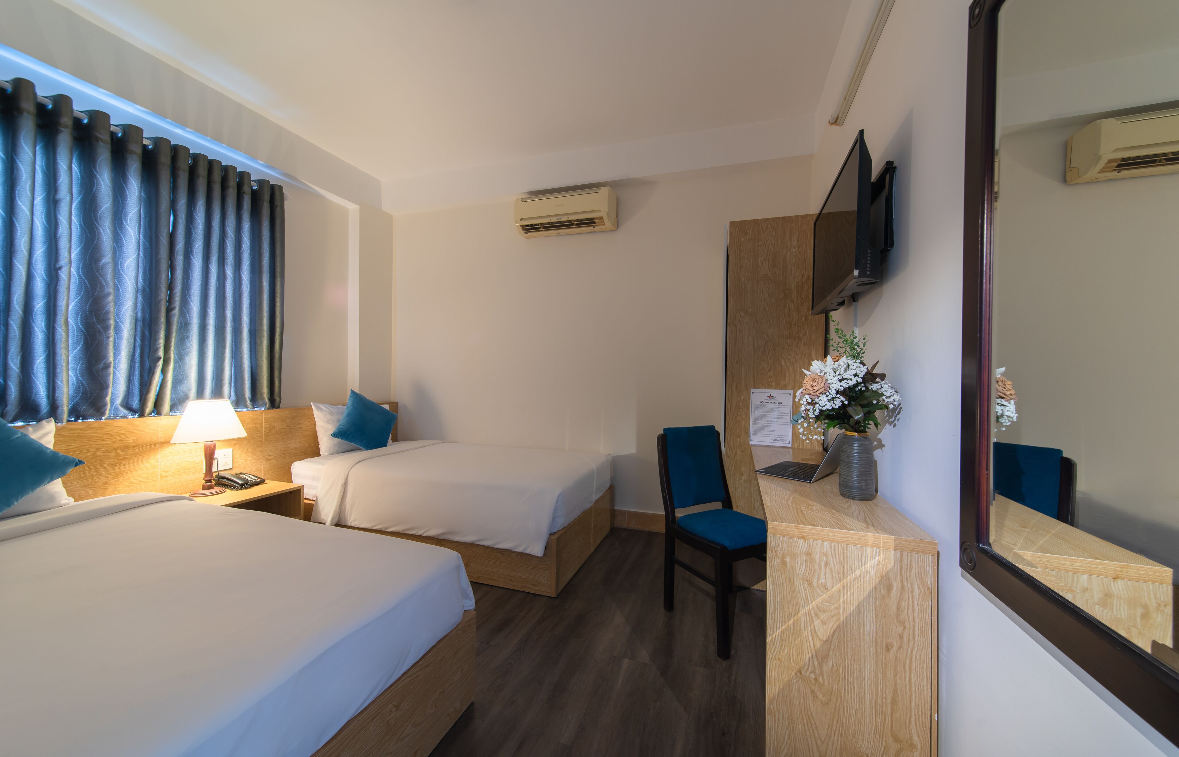 A hotel room with two beds, a desk with a chair, a TV mounted on the wall, and a window with curtains.