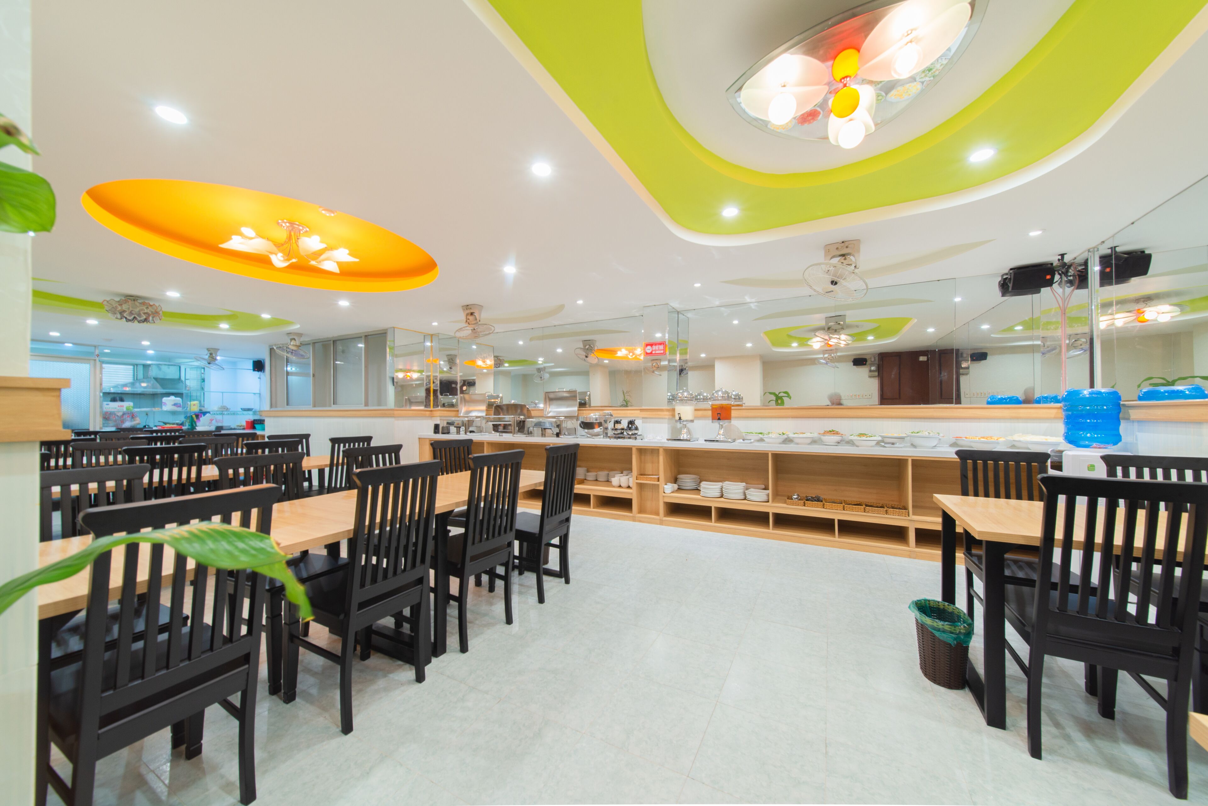 Un espace repas moderne avec des tables et des chaises en bois, un comptoir central doté d’un distributeur d’eau et un plafond agrémenté de touches de vert et de blanc.