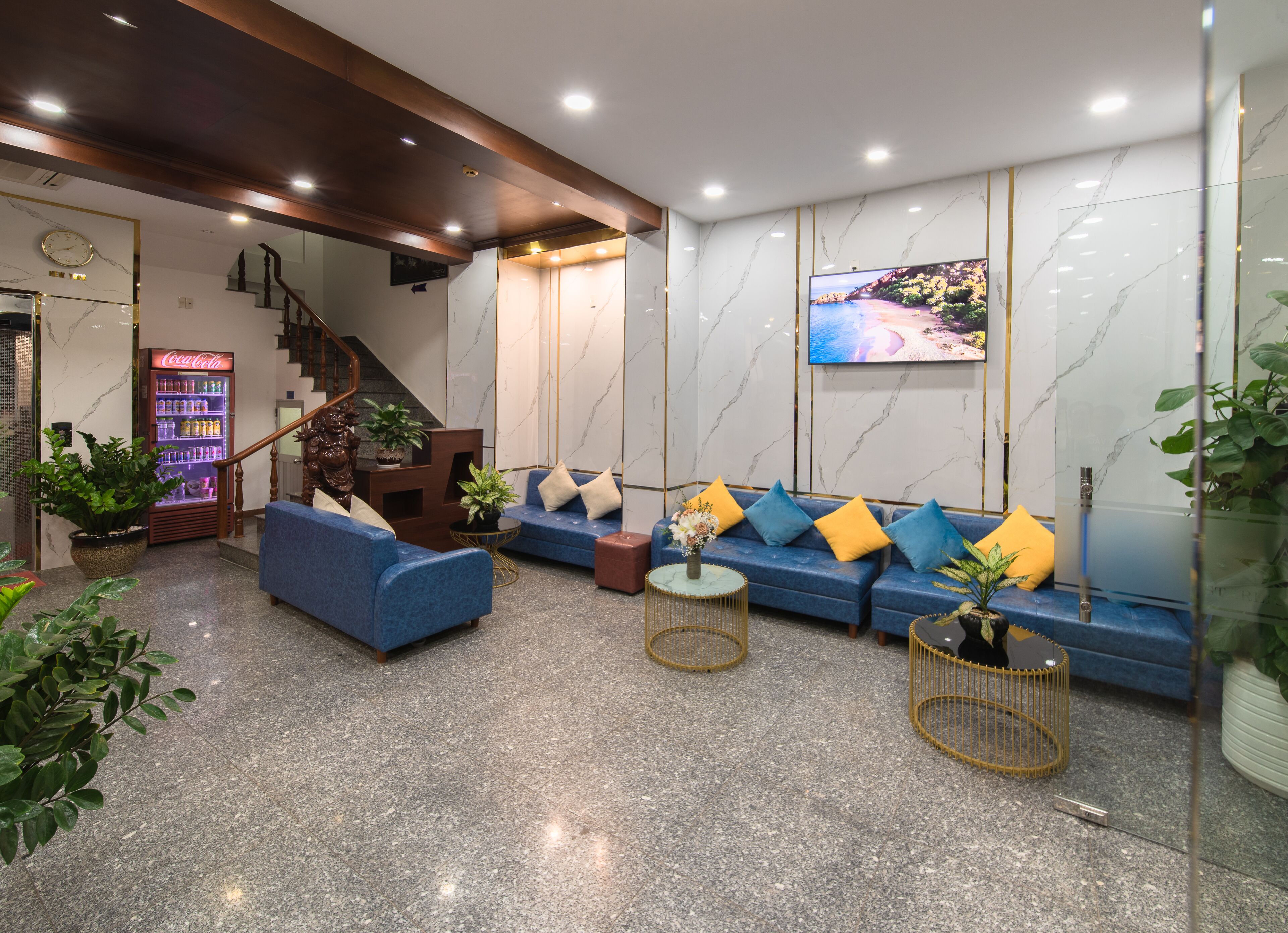 A modern lobby with a blue sofa set, a Coca-Cola refrigerator, a staircase, and a TV on the wall.
