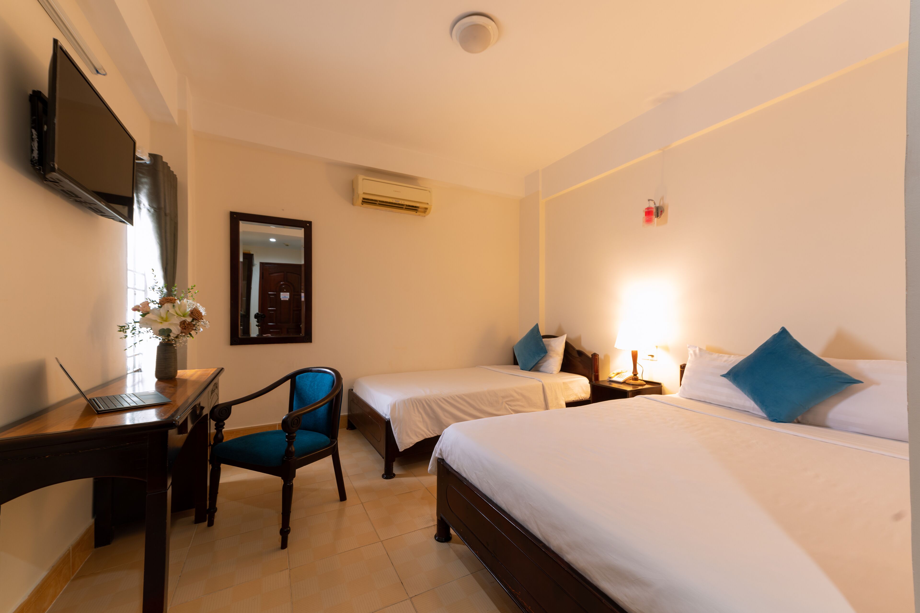 A hotel room with two single beds, a desk with a laptop, a chair, a TV mounted on the wall, and an air conditioning unit.