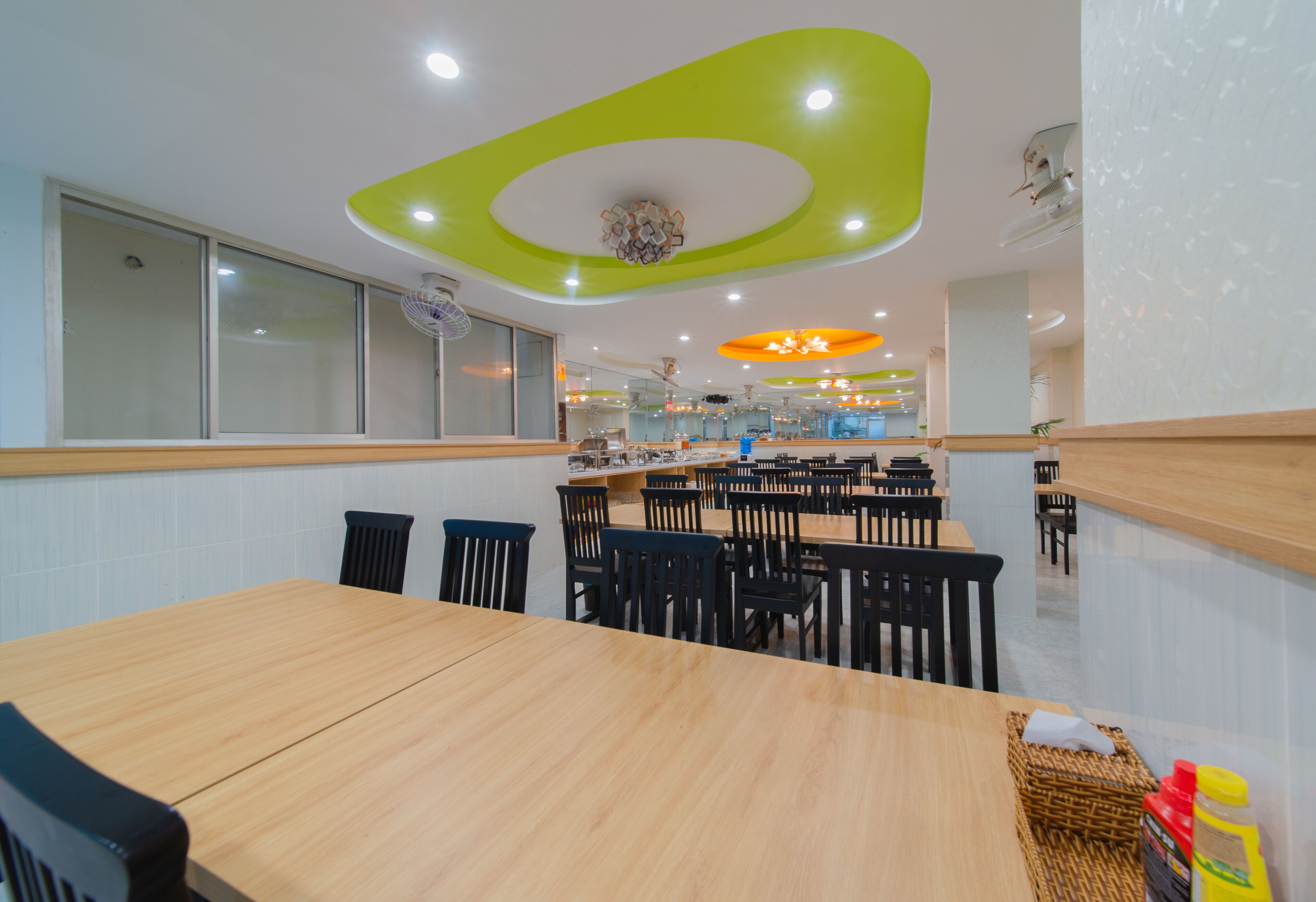 A modern dining area with wooden tables and black chairs, a green ceiling design, and a central light fixture.