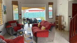 Un salon spacieux avec un canapé gris, un fauteuil rouge et une petite table ronde. Une grande fenêtre offre une vue sur un immeuble rouge situé à l’extérieur.