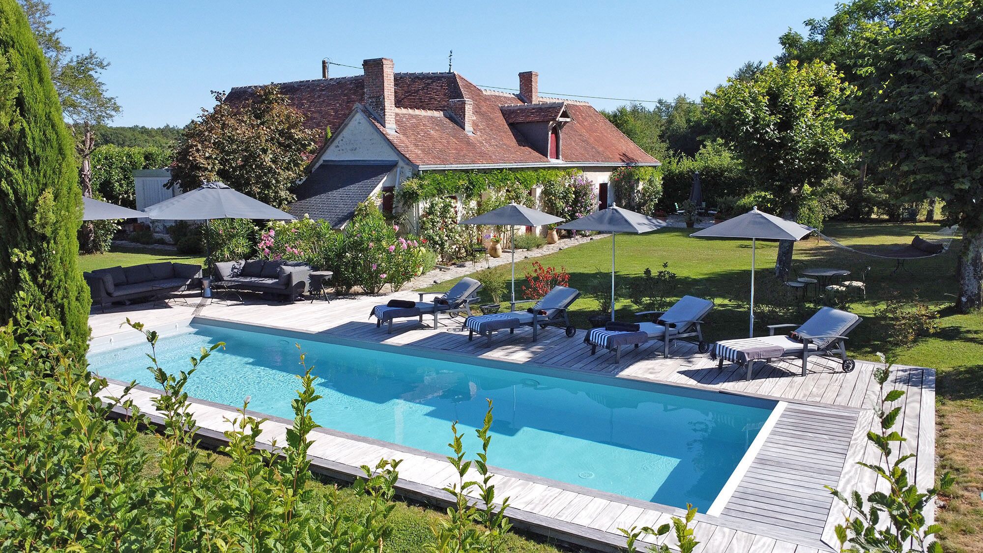 Een zwembad met ligbedden en parasols, omringd door een tuin en een huis.