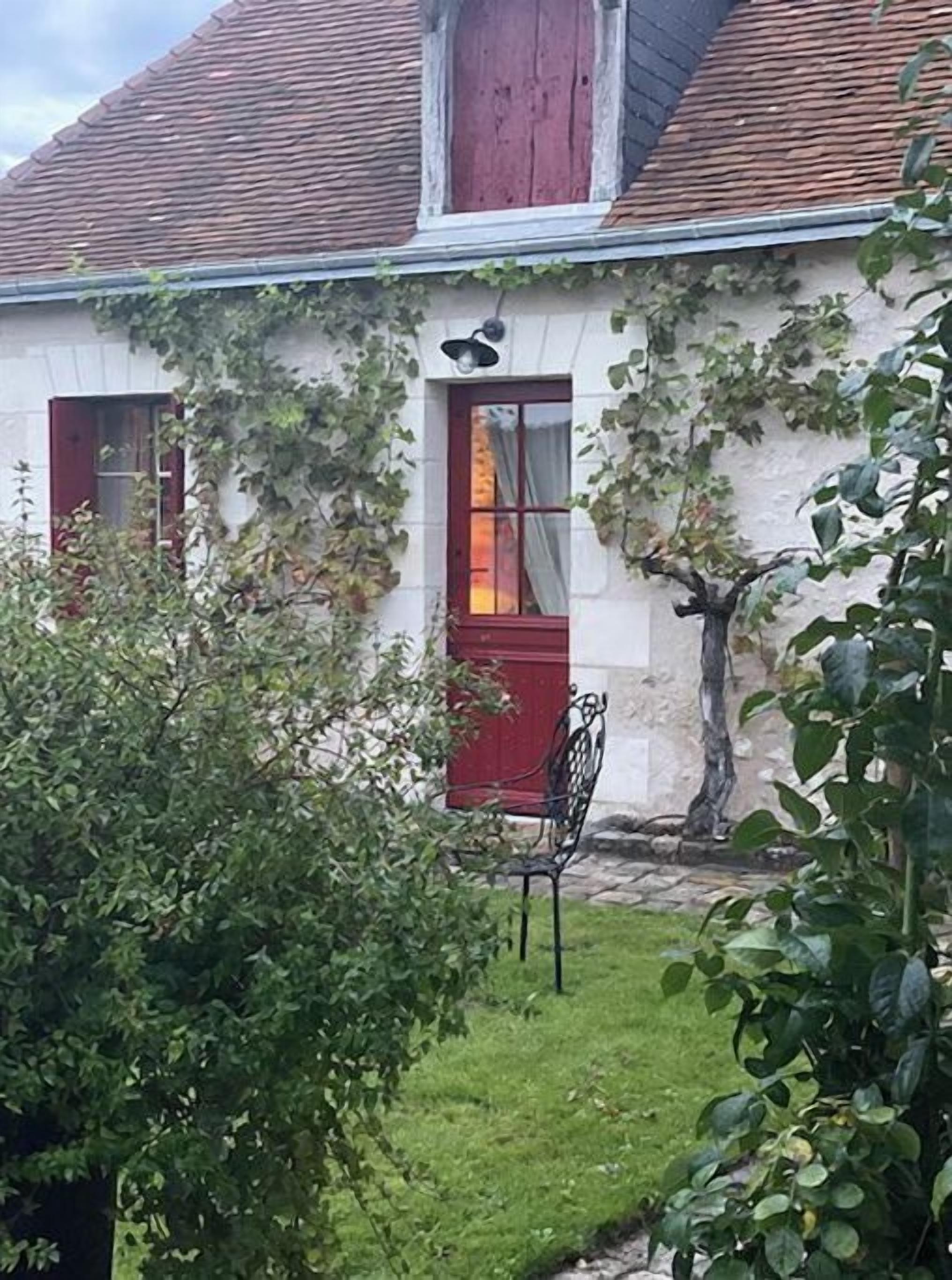 A white building with a red door and window shutters, a small garden with a metal chair, and a tree.