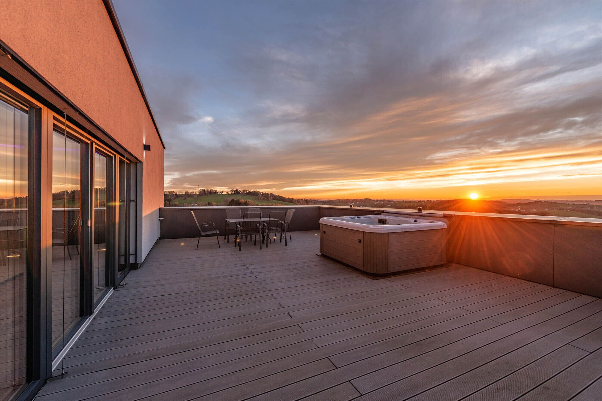 Een houten terras met een jacuzzi, stoelen en een tafel, met uitzicht op een zonsondergang.