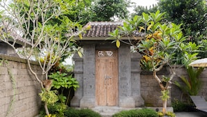 Une porte en bois traditionnelle surmontée d’un toit en tuiles, entourée d’un mur de pierre et d’une végétation luxuriante.