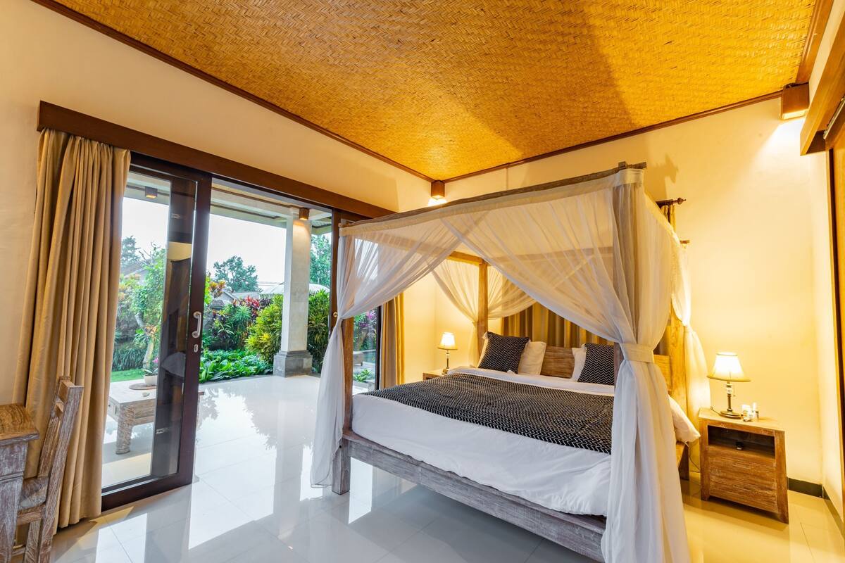 A spacious bedroom with a large bed, a nightstand, and a view of a garden through a sliding glass door.
