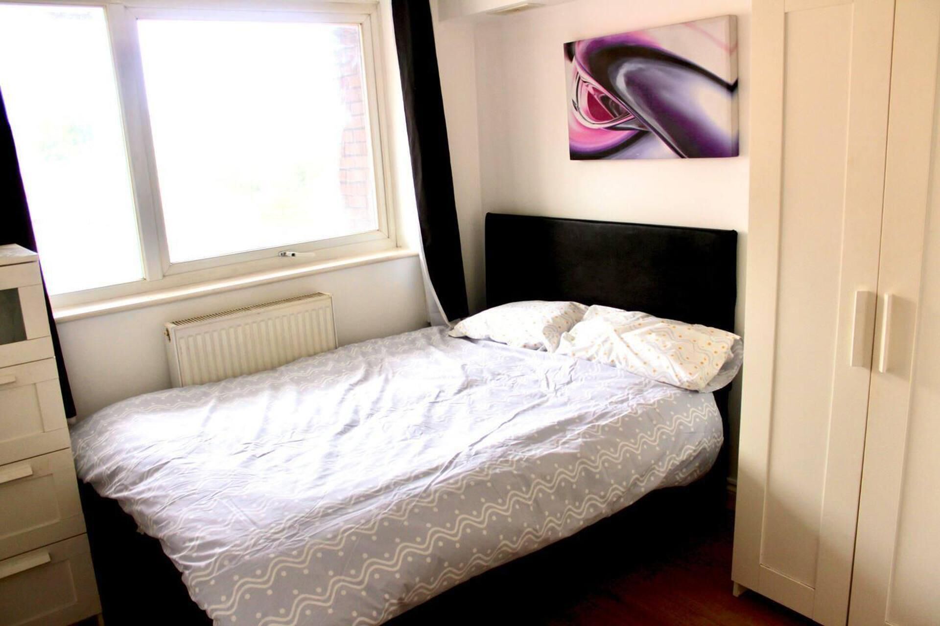 A bedroom with a bed, a radiator, a window, a wardrobe, and a painting on the wall.