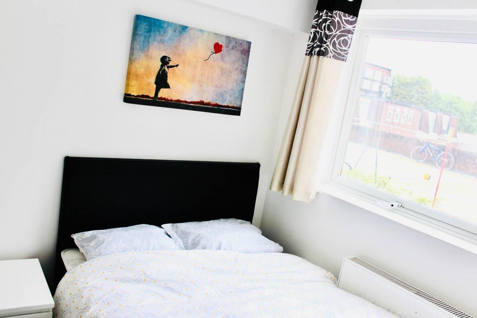 A bedroom with a bed, a nightstand, a window with curtains, and a wall-mounted painting.