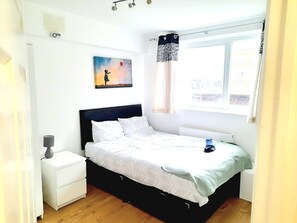 Ein Schlafzimmer mit einem Bett, einem Nachttisch, einem Wandheizkörper, einem Fenster mit Vorhängen und einem Bild an der Wand.