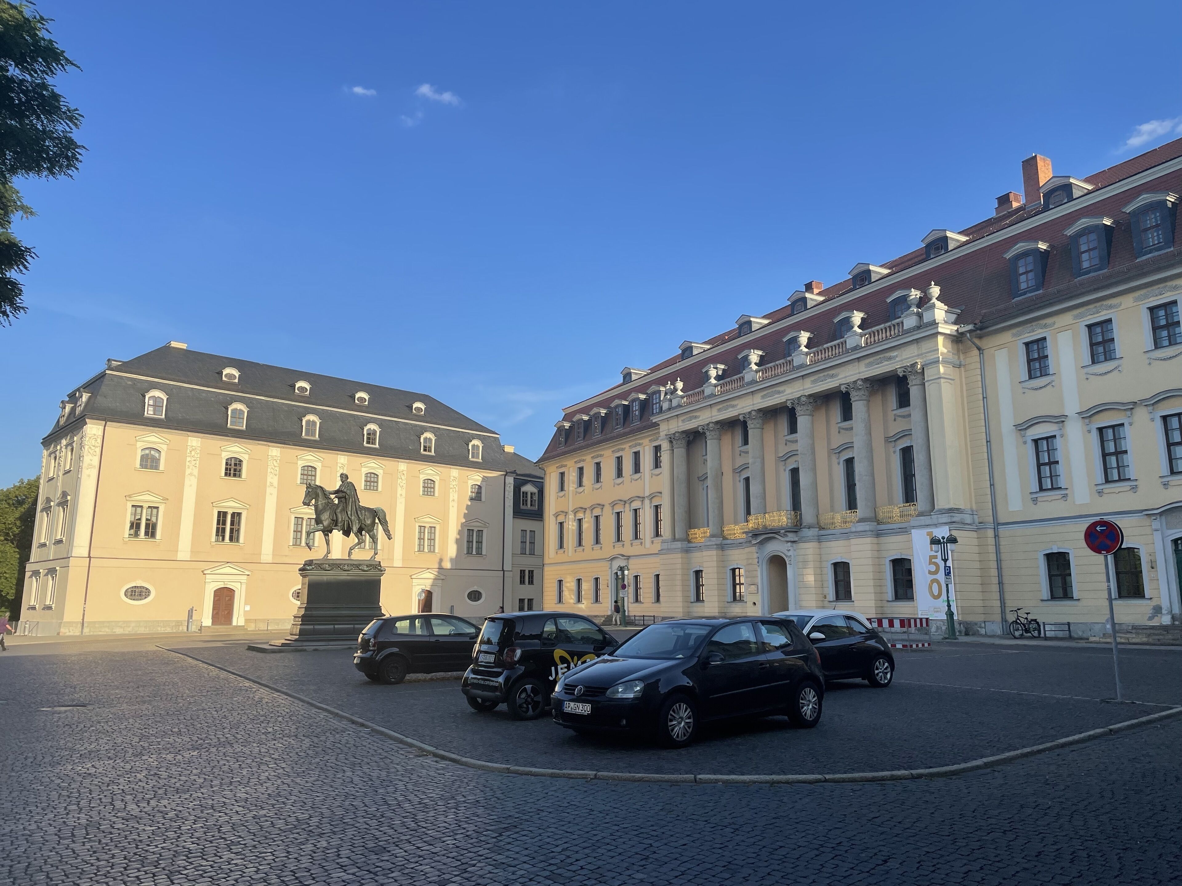 A historic building with classical architecture, a statue of a man on horseback, and a cobblestone street with parked cars.