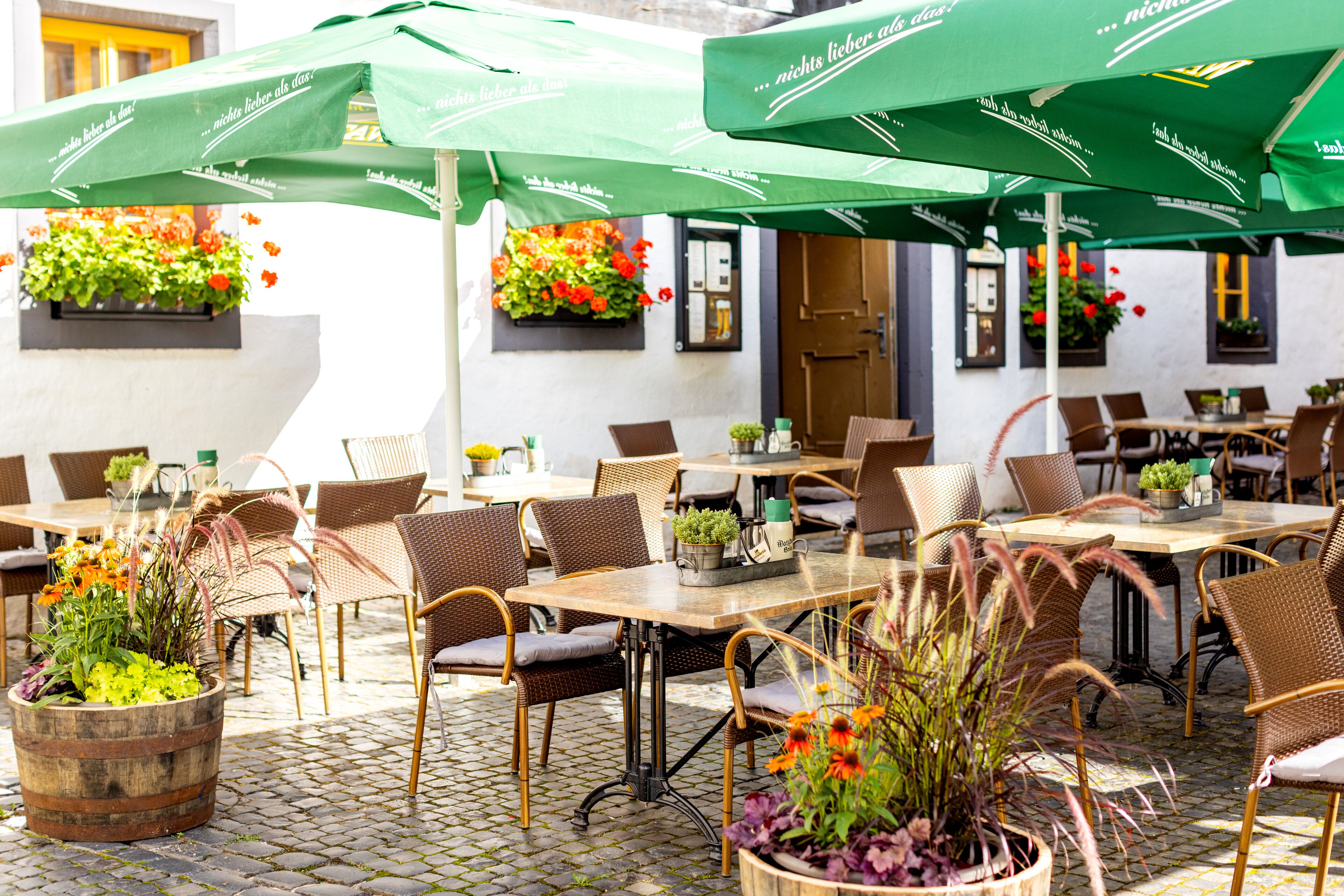 Un café al aire libre con sillas y mesas de mimbre, sombrillas verdes y macetas con flores en las paredes.