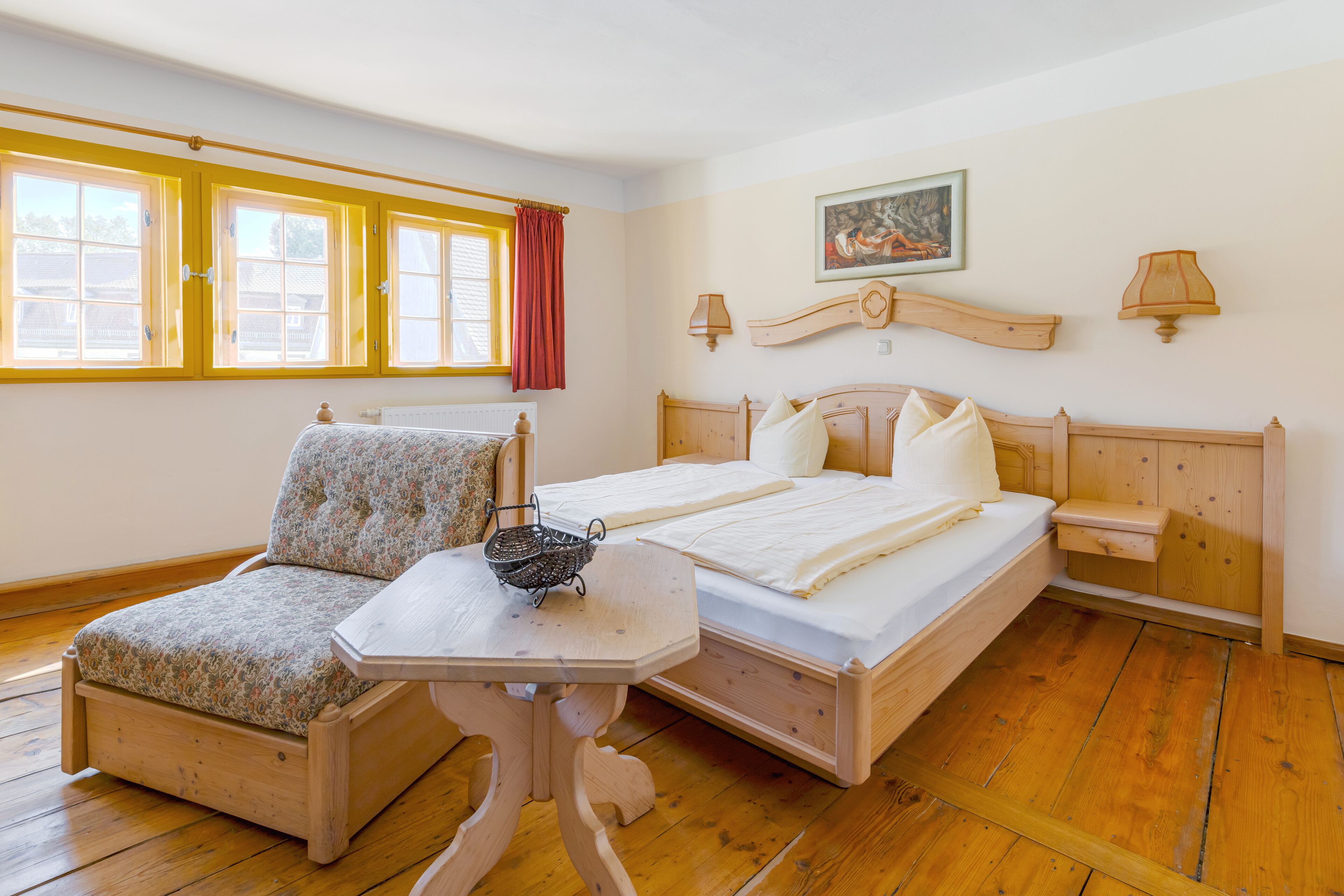 Ein Schlafzimmer mit Holzboden, einem Bett mit Holzüberbau, einem Sessel mit Blumenkissen, einem kleinen Tisch und zwei Wandlampen.