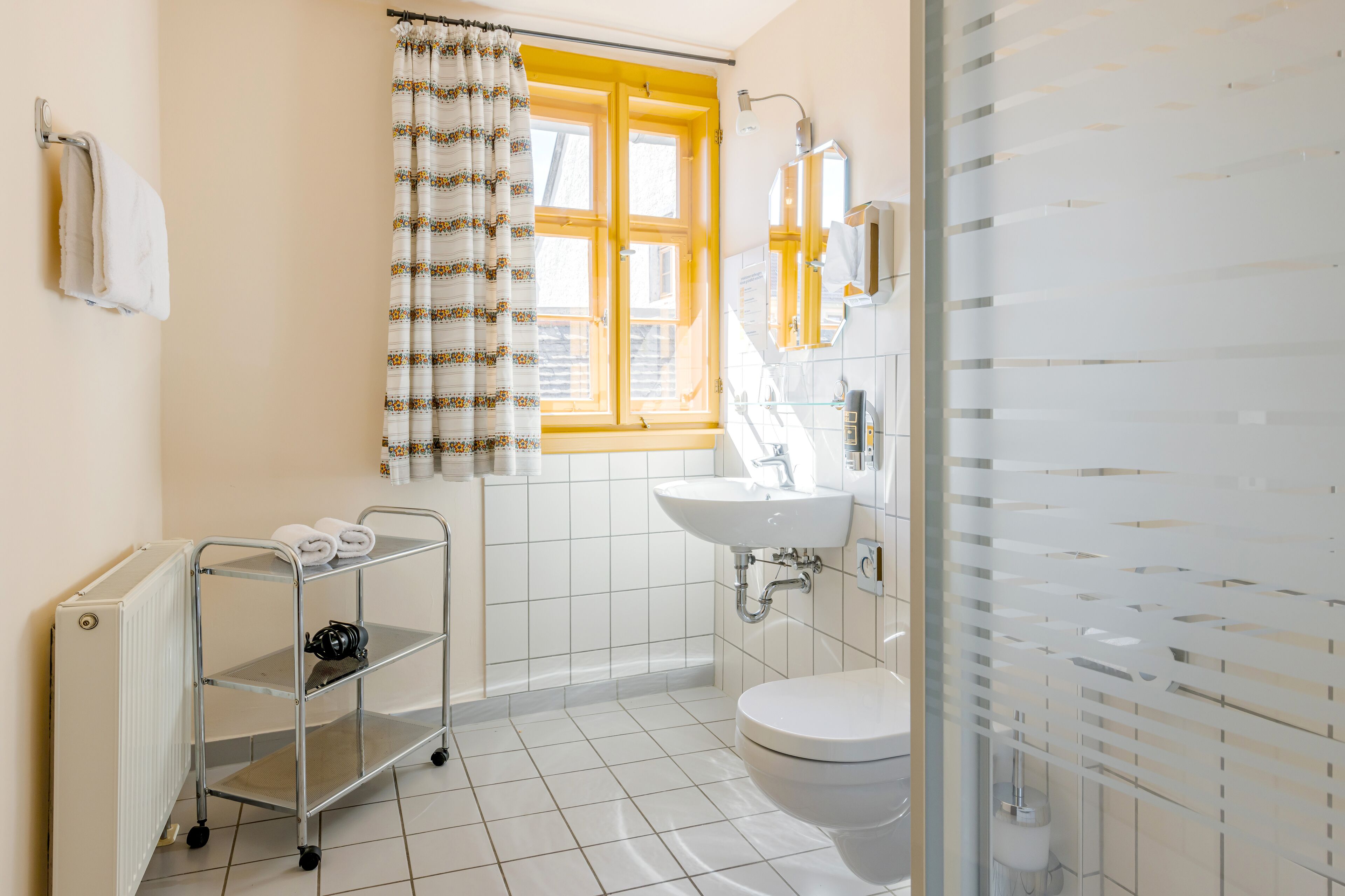 Un baño con inodoro blanco, lavamanos, espejo, radiador y repisa con toallas.