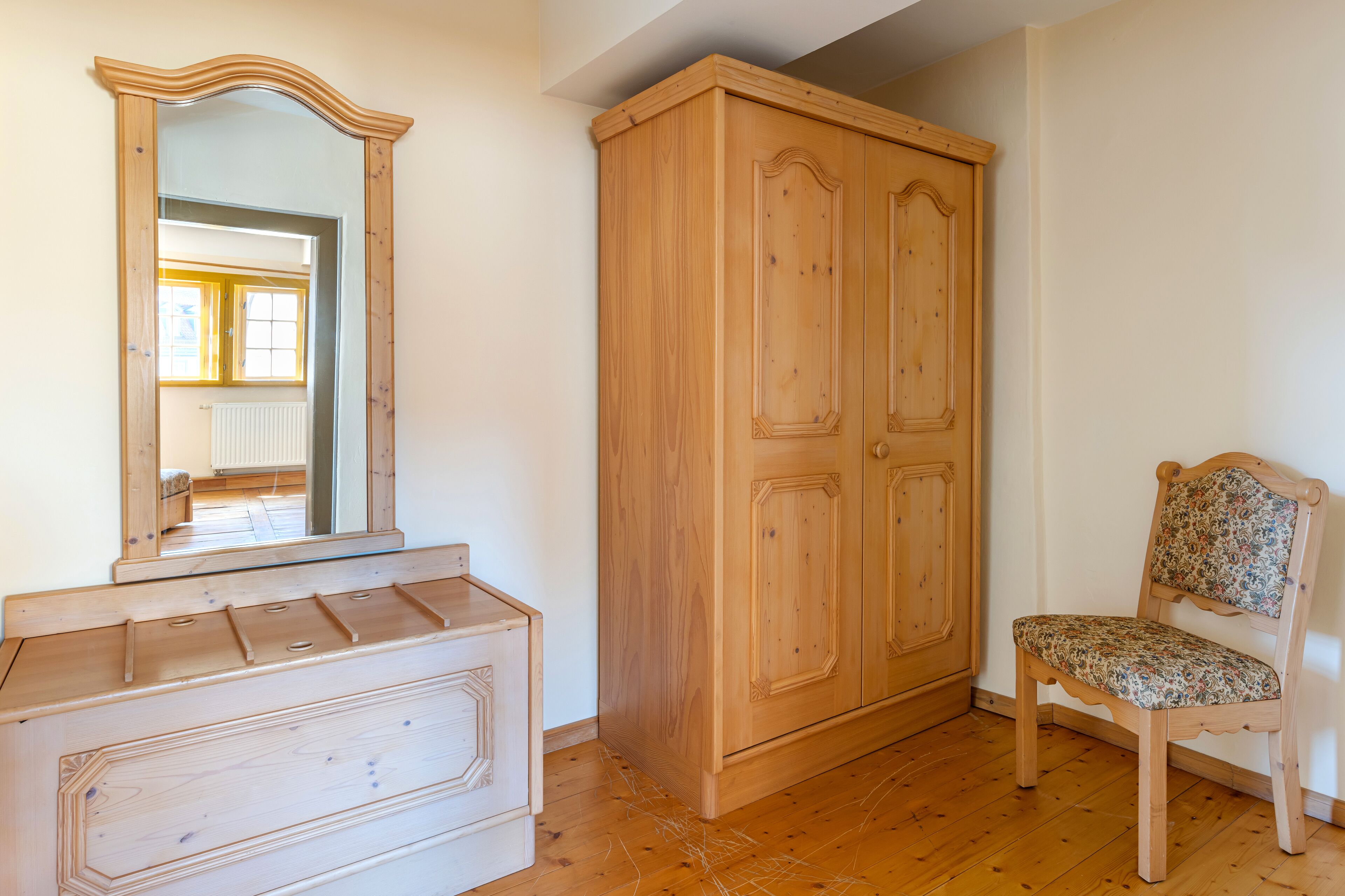 A wooden wardrobe, a mirror, and a chair in a room with wooden flooring.