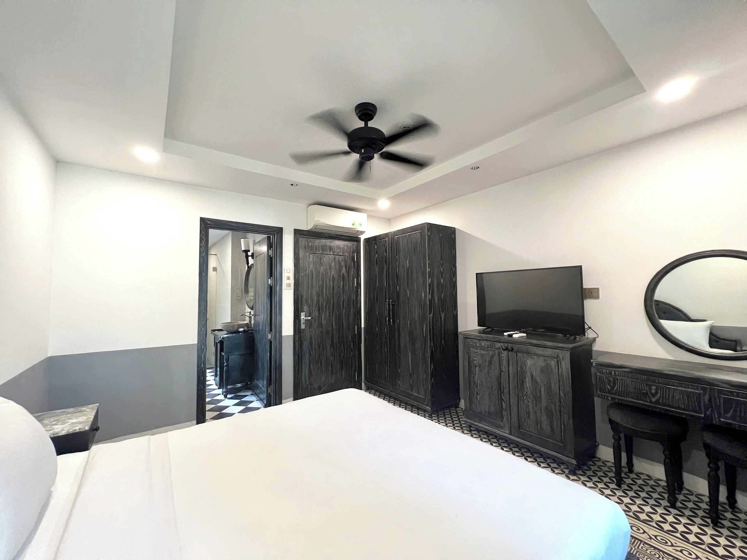 A hotel room with a bed, a desk with a TV, a wardrobe, a ceiling fan, and a bathroom visible through an open door.