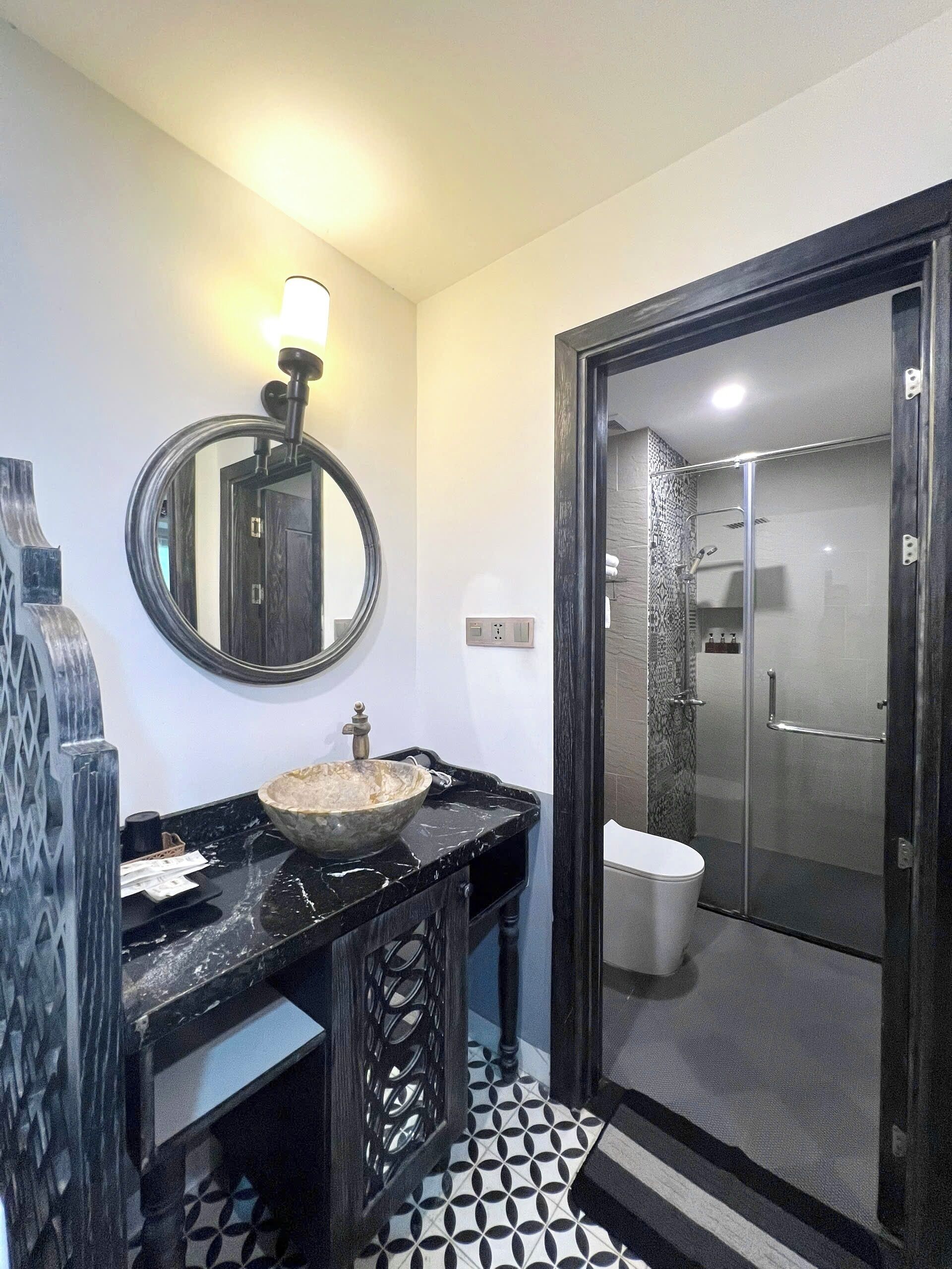 A bathroom with a round mirror, a vessel sink, a toilet, and a glass-enclosed shower.