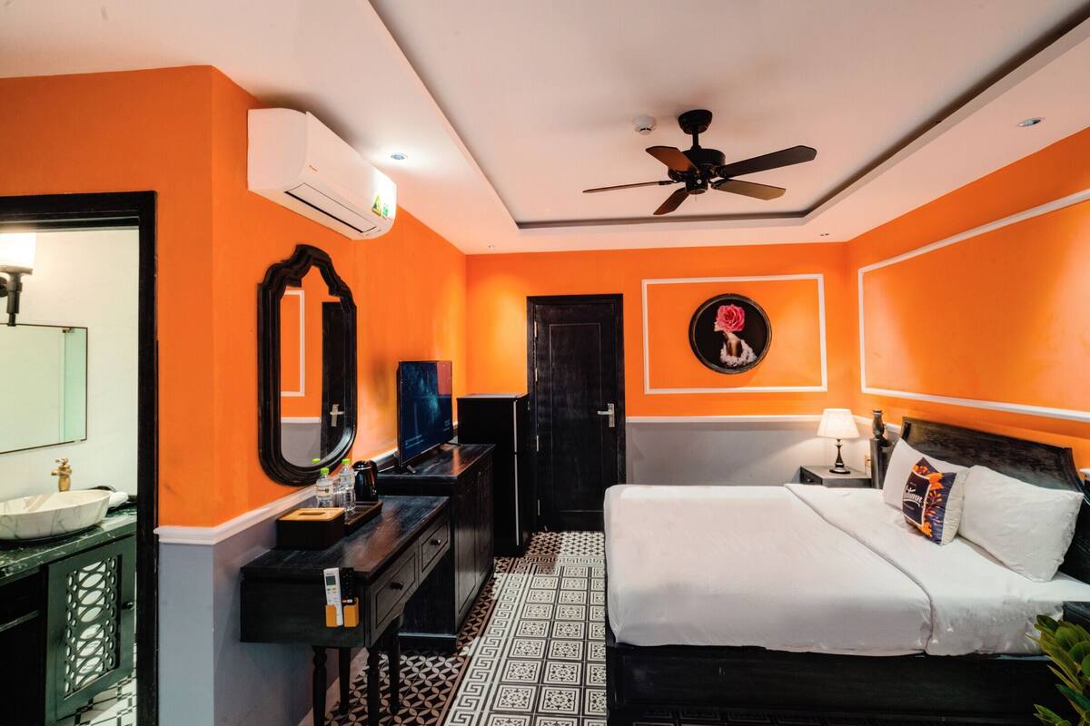 A hotel room with an orange wall, a ceiling fan, a bed with white bedding, a TV, a mirror, and a bathroom with a sink and toilet.