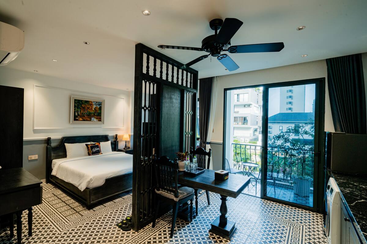 A hotel room with a bed, a desk, a chair, a ceiling fan, and a balcony with a view of buildings.