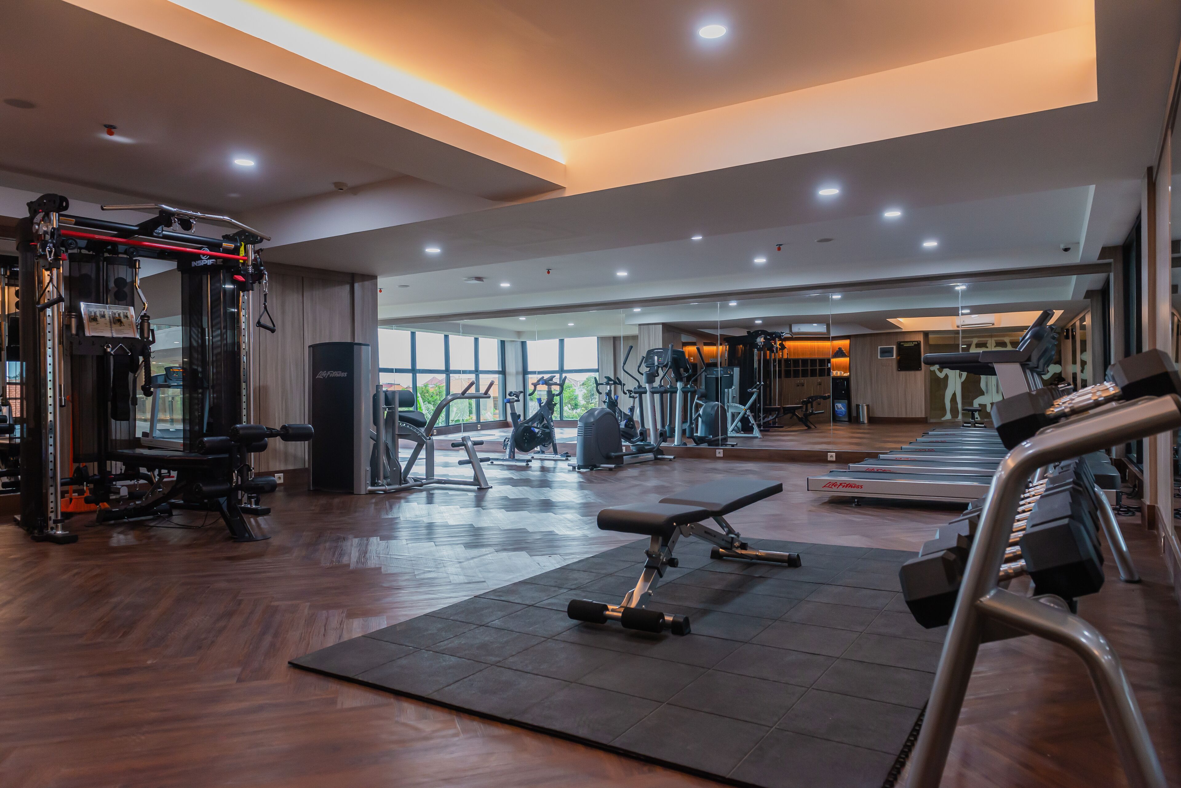Un gimnasio amplio con diferentes equipos de ejercicio, ventanas grandes y piso de madera.