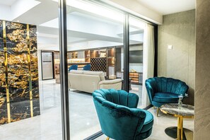 A modern interior with a glass partition, teal armchairs, a marble wall, and a dining area in the background.