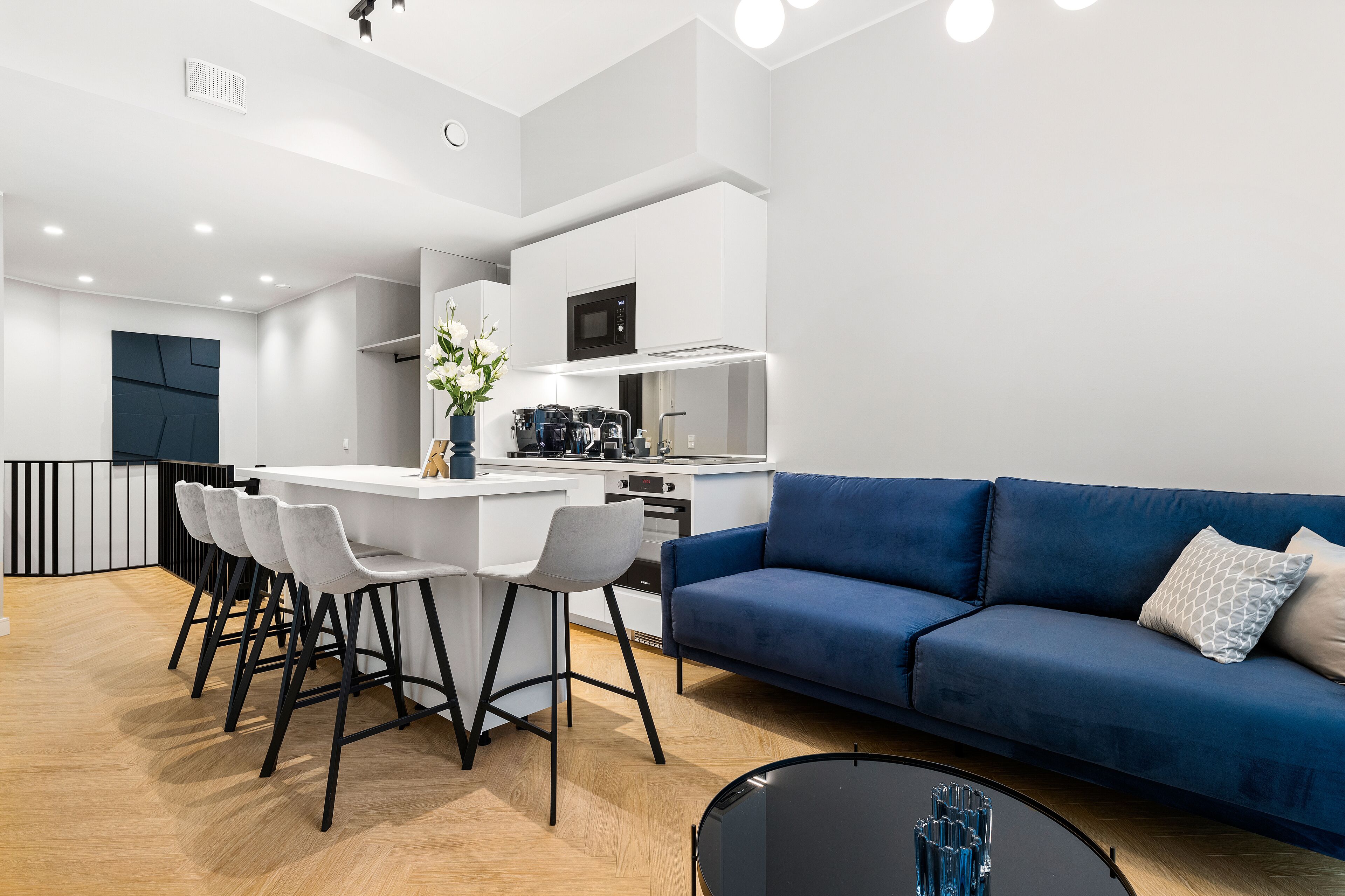 A modern kitchen with white cabinets, a black microwave, and a coffee machine. There is a blue sofa with a patterned pillow and a small table with decorative items.