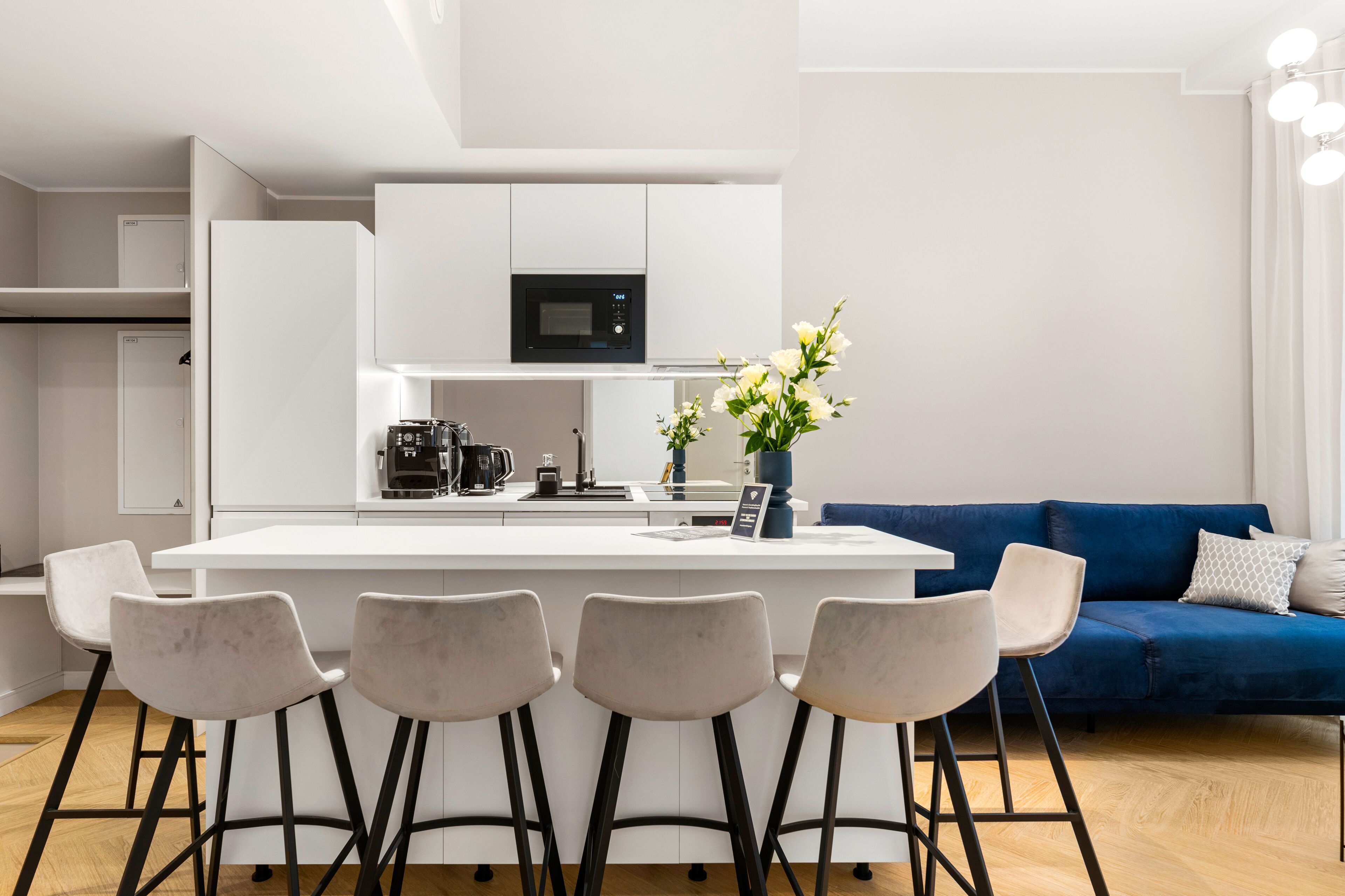Eine moderne Küche mit weißen Schränken, einer schwarzen Mikrowelle und einer Kaffeemaschine. Vier Barhocker und ein blaues Sofa mit weißen Kissen sind ebenfalls vorhanden.