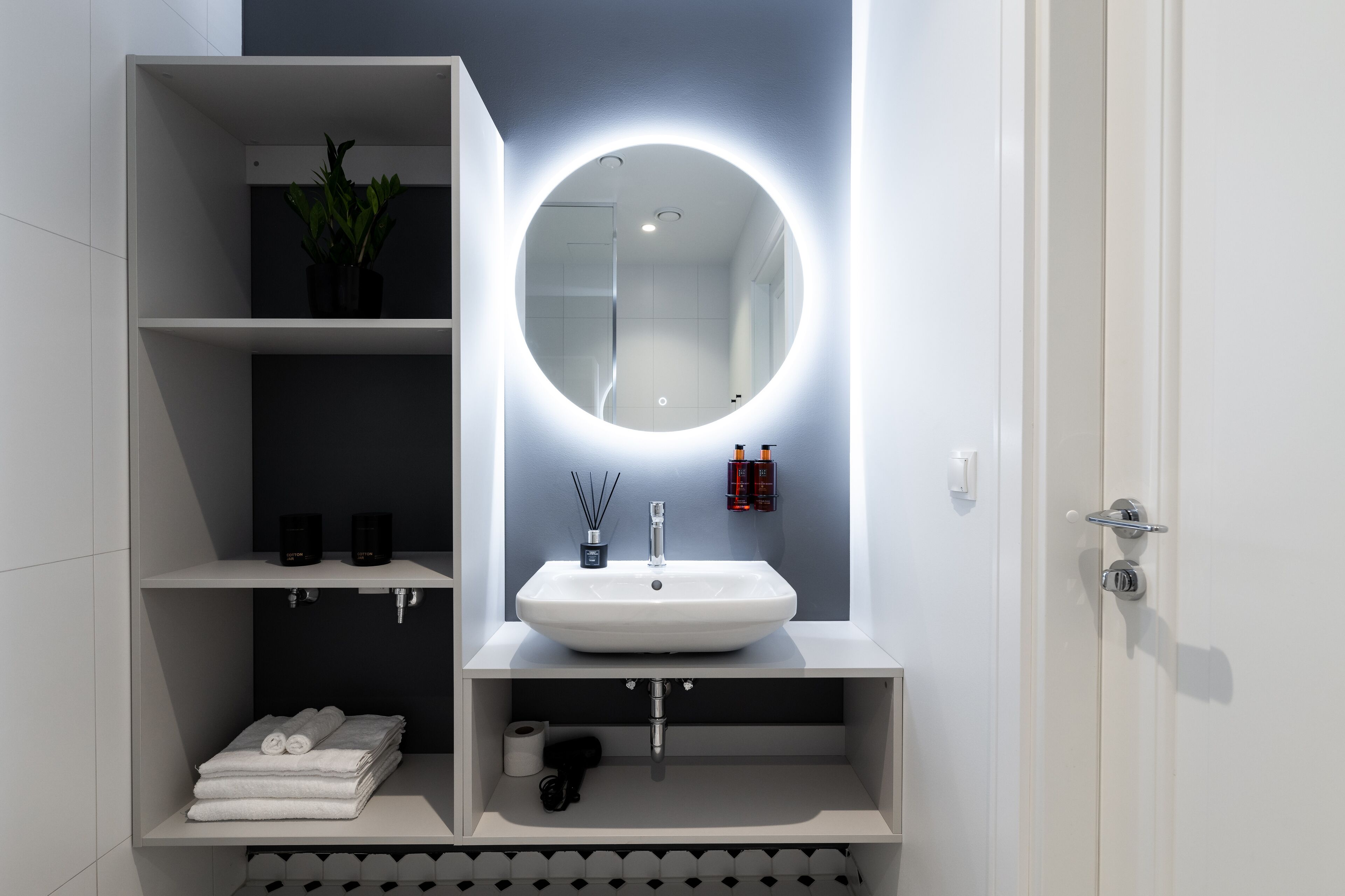 A modern bathroom with a white sink, a round mirror with underglow lighting, and shelves with toiletries.