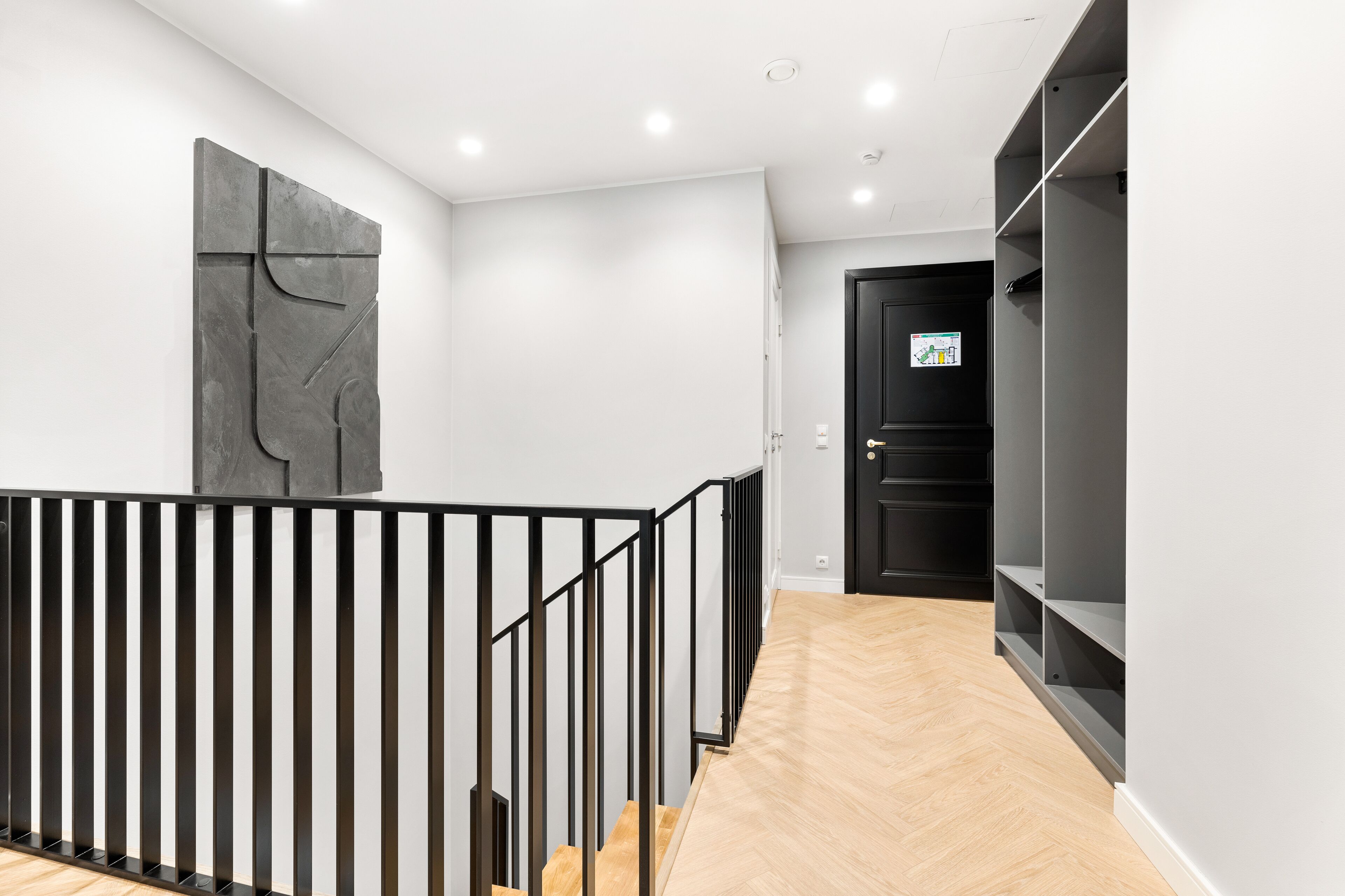 Ein modernes Interieur mit einer Treppe, einer schwarzen Tür mit einem Schild und einer grauen Wand mit einem geometrischen Kunstwerk.