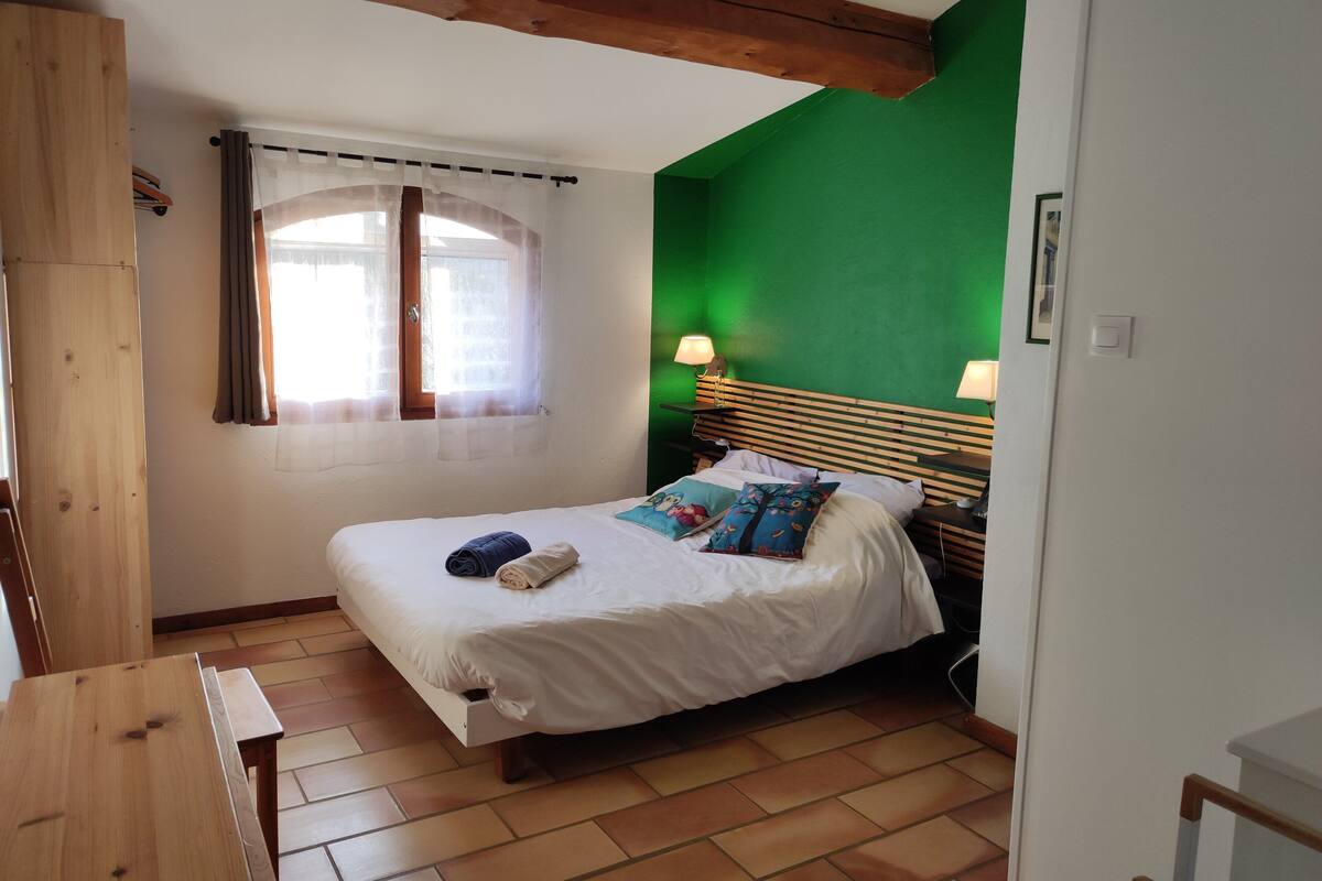 A bedroom with a wooden bed frame, a green accent wall, a window with sheer curtains, and two bedside lamps.