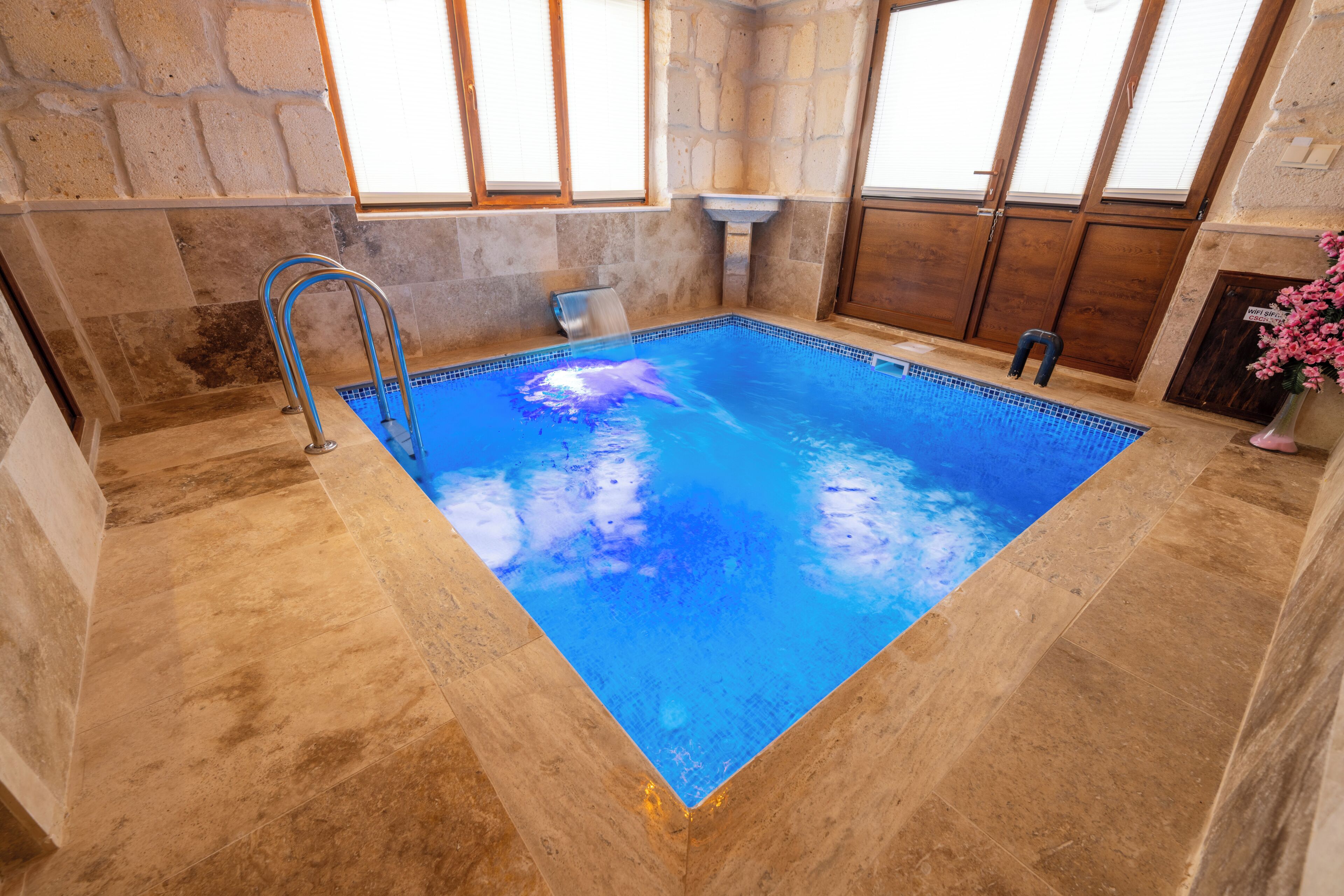 An indoor swimming pool with stone walls, a metal ladder, and a window with blinds.