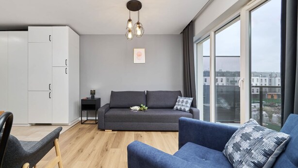 A modern living room with a grey sofa, a blue armchair, a white cabinet, and a large window offering a city view. - Modern Studio on Jagodno | Balcony and Parking (Wrocław)
