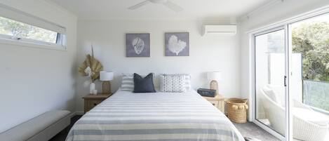 A bedroom with a large bed, a ceiling fan, two wall art pieces, a nightstand with a lamp, and a view through a sliding glass door.