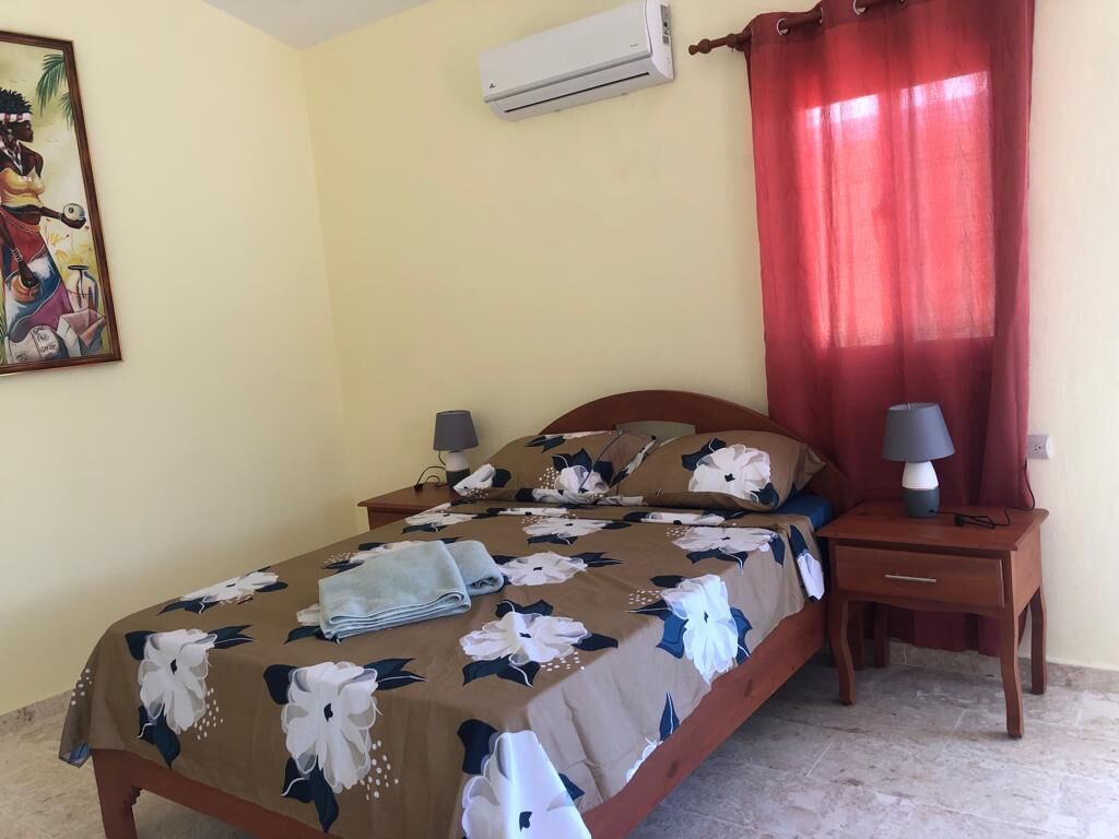 Een slaapkamer met een bed, nachtkastjes, een airconditioner en een raam met rode gordijnen.