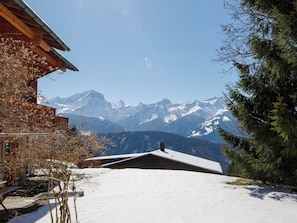 A wooden cabin with a snow-covered roof, a clear view of snow-capped mountains, and a pine tree to the right. - Chaperon Rouge A3 by Interhome (Villars)