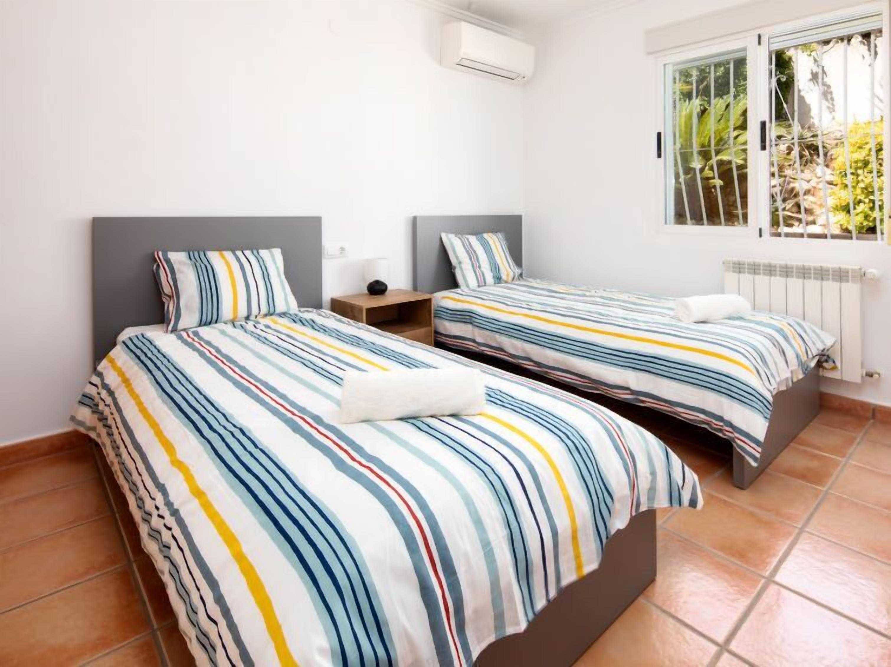 A bedroom with two beds, each with striped bedding, a nightstand, and a window with a view of greenery.