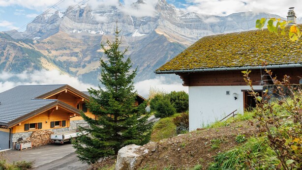 Una cabaña de montaña con fachada de madera y techo inclinado, rodeada de exuberante vegetación y un telón de fondo de picos nevados.