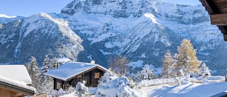 Una cabaña de madera con techo nevado, rodeada de árboles cubiertos de nieve y montañas.