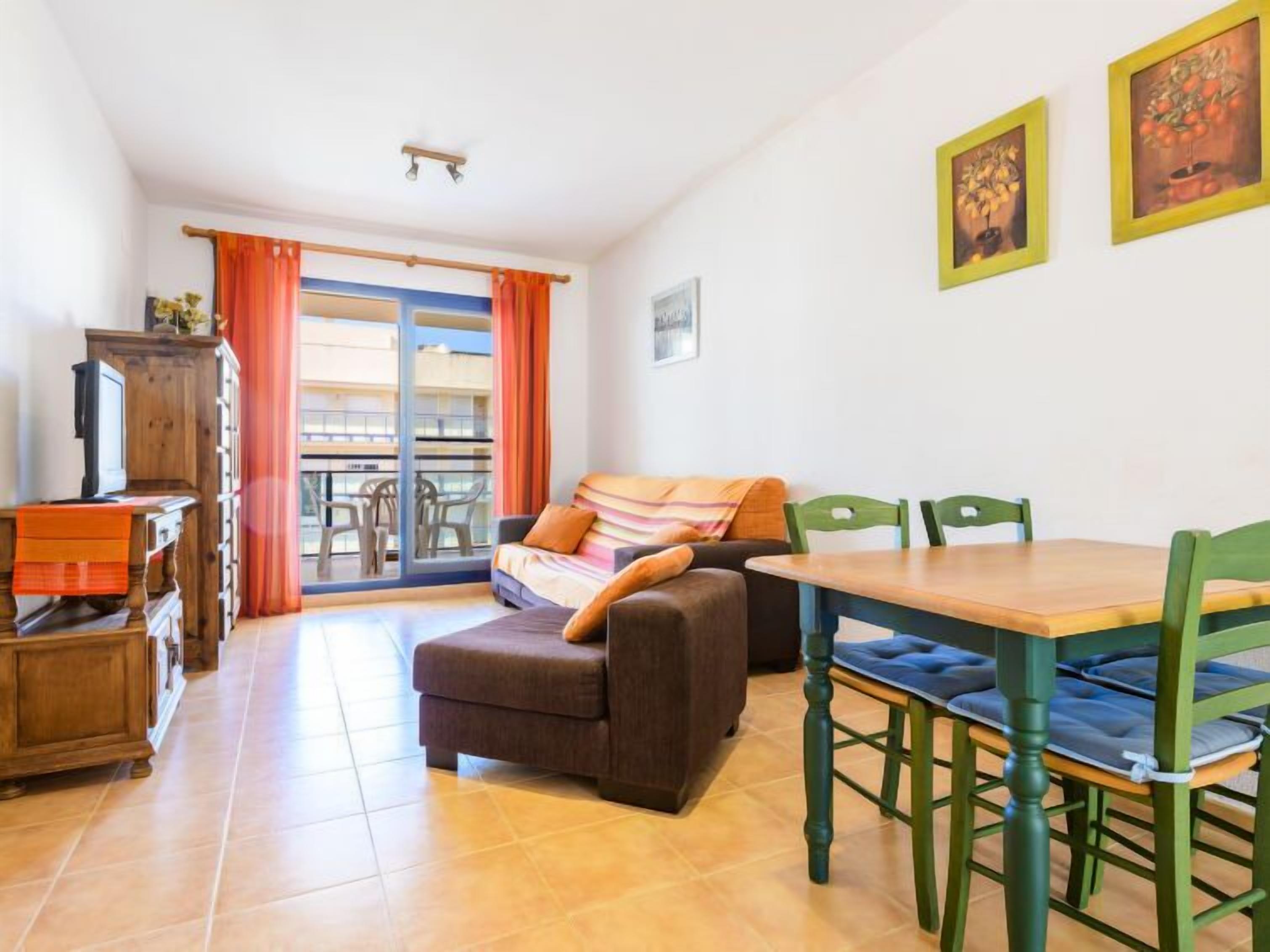 A cozy living room with a sofa, a wooden table, and chairs. There is a balcony with orange curtains and a view of buildings.