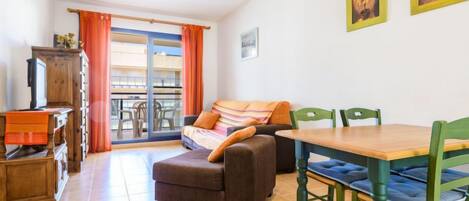 A cozy living room with a sofa, a wooden table, and chairs. There is a balcony with orange curtains and a view of buildings.