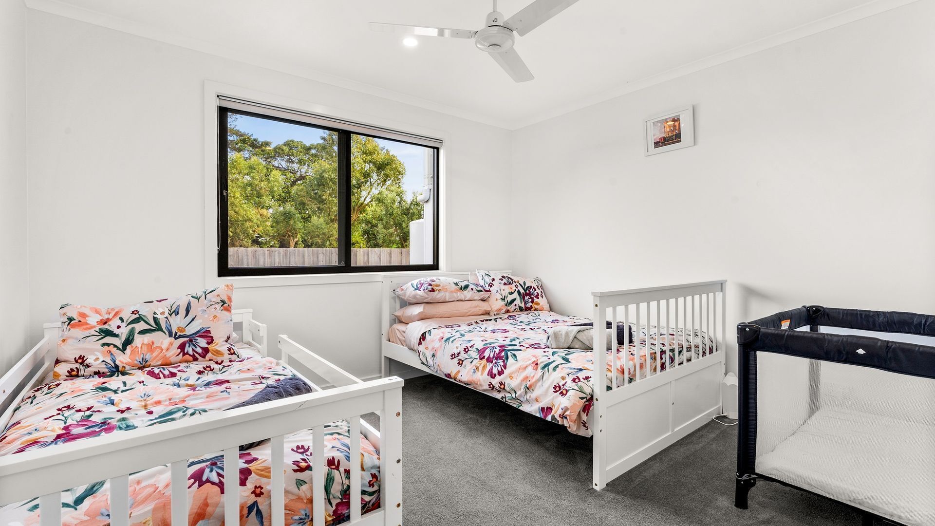 A bedroom with two single beds, a crib, a ceiling fan, a window with a view of trees, and a framed picture on the wall.