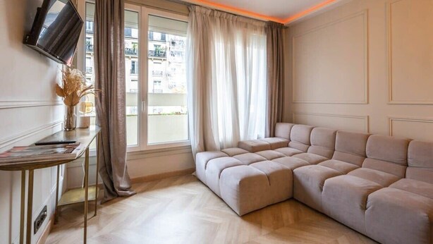 A modern living room with a sectional sofa, a flat-screen TV mounted on the wall, and a large window with curtains. - Petit Violette I Amazing 1 Bed Apt in Paris (Paris)