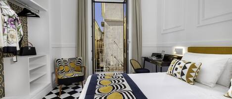 Une chambre d’hôtel avec un sol à carreaux, un lit agrémenté d’oreillers à motifs, un bureau avec un ordinateur portable, et une vue sur l’extérieur par une porte ouverte.