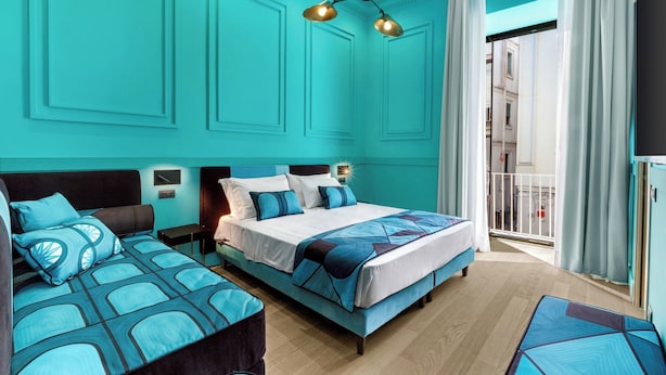A hotel room with a teal color scheme, a large bed, two smaller beds, a nightstand, a lamp, and a view of the outside. - Maison Iovino Luxury Rooms (Sorrento)