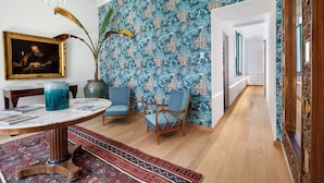 A room with a patterned wallpaper, a table with magazines, a chair, a vase, and a chandelier. - Maison Iovino Luxury Rooms (Sorrento)