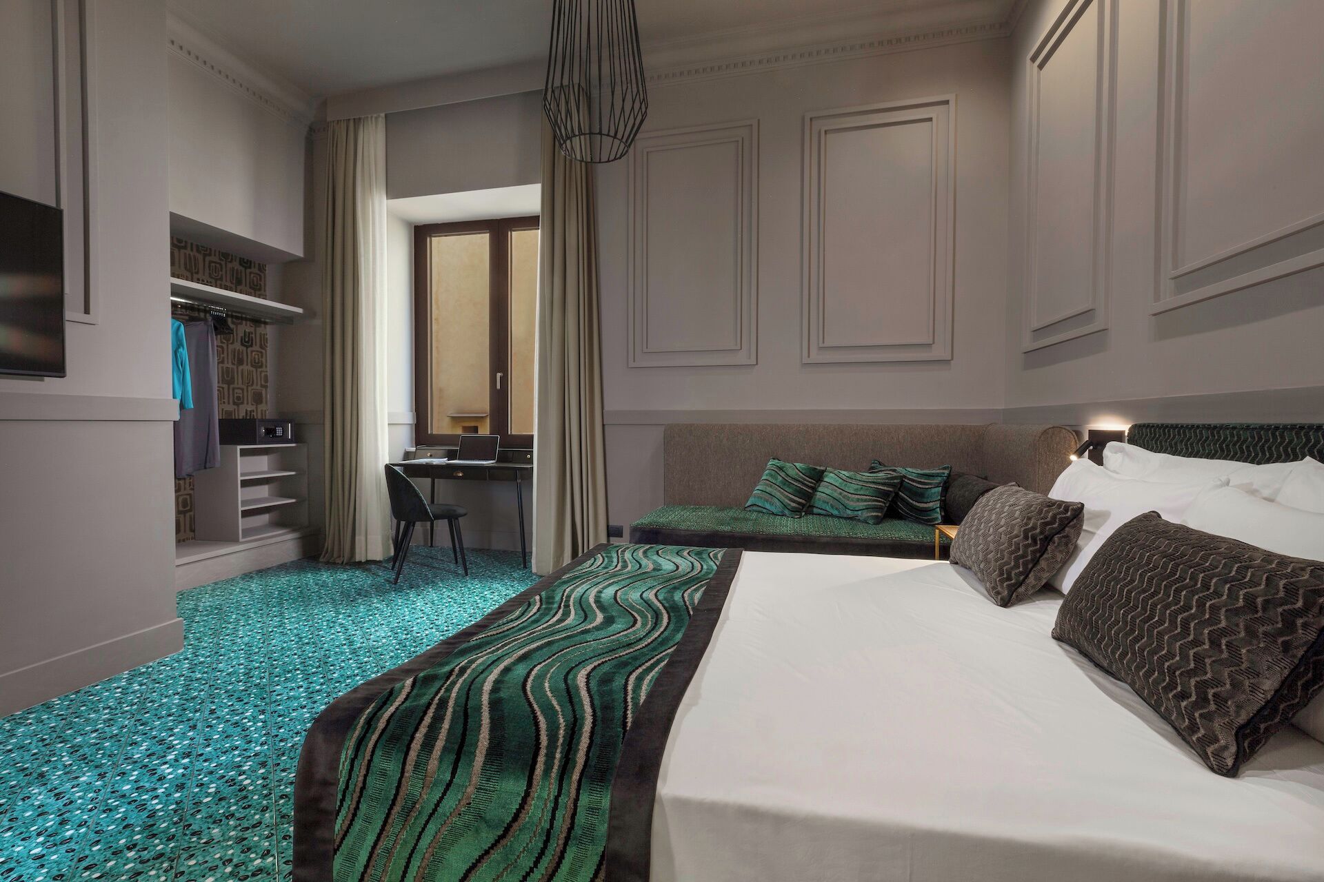 A hotel room with a bed, a desk, and a chair. There is a wardrobe and a mirror.
