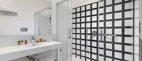 Uma casa de banho moderna com cabine de duche em vidro, lavatório branco com artigos de higiene pessoal e um espelho grande.