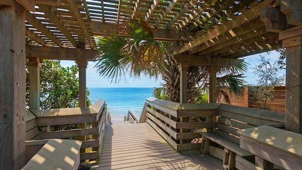 Una terraza de madera que da a una playa, con una pérgola de madera sobre ella.