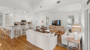 A spacious living area with a sofa, chairs, a coffee table, and a TV. There is a kitchen with white cabinets and a dining area with bar stools. - Near to beach & close to the pool, Seagrove + Free Attraction Tickets! (Santa Rosa Beach)